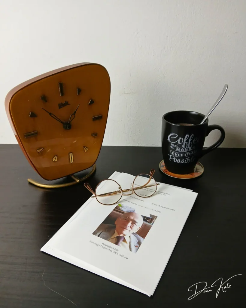 A desk with a clock, a pair of eyeglasses atop documents, and a coffee mug with motivational text sits on a coaster—as if inviting writing through grief or crafting a bilingual elegy in the quiet morning light.