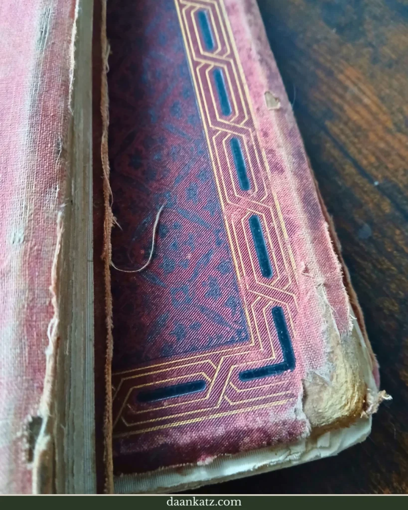 A close-up of a worn, antique book cover with frayed edges and a geometric gold design—resting on a wooden surface, it bears visible traces of forgotten times.