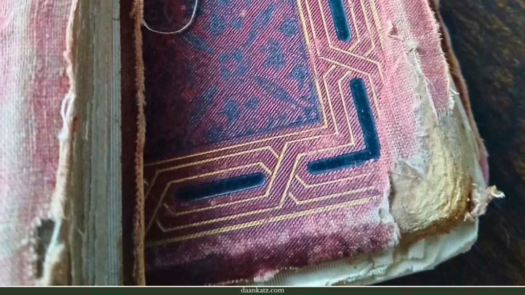A close-up of a worn, antique book cover with frayed edges and a geometric gold design—resting on a wooden surface, it bears visible traces of forgotten times.