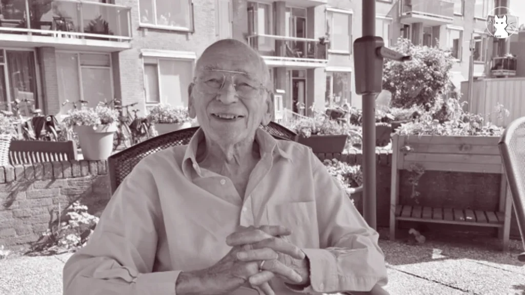 An elderly man wearing glasses sits outside in a garden, smiling at the camera. He is seated at a table with plants, bikes, and apartment buildings in the background.